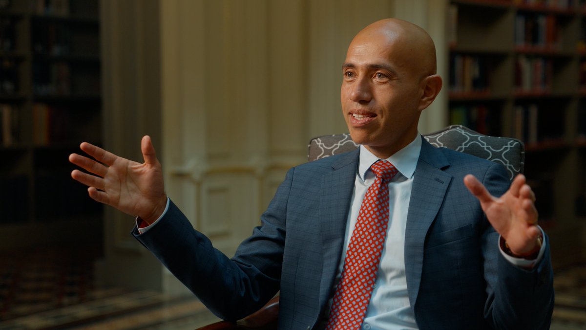 This is a professional photograph showing a bald man in a business suit during what appears to be an interview or speaking engagement. He is wearing a dark blue/navy suit with a red patterned tie and white dress shirt, seated in an ornate chair with decorative upholstery. His hands are raised in an expressive gesture, suggesting he is mid-conversation or making a point. The setting appears to be an elegant interior space with bookshelves visible in the blurred background, giving it a library ...