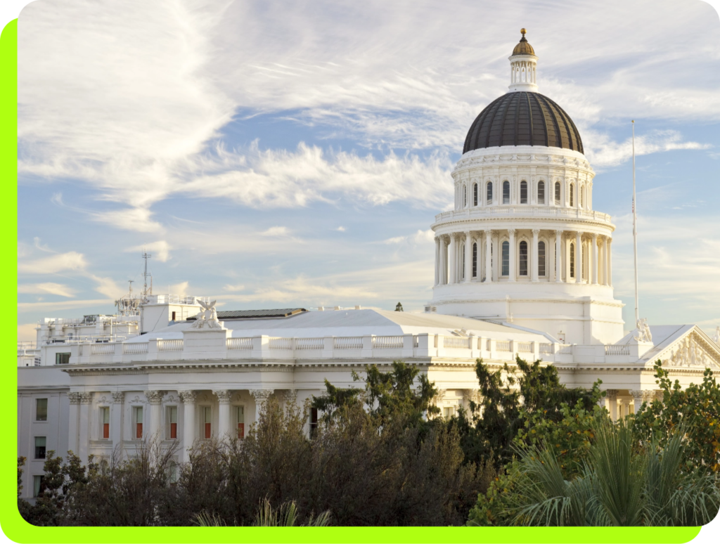 This is a photograph showing the California State Capitol building in Sacramento, featuring its distinctive white neoclassical architecture with a prominent dome topped by a dark cupola and golden finial. The image captures the building from a distance, showing both the domed rotunda and the lower legislative wings with their classical columns and white facades. The scene is framed by lush vegetation including palm trees and other greenery in the foreground, emphasizing California's Mediterra...