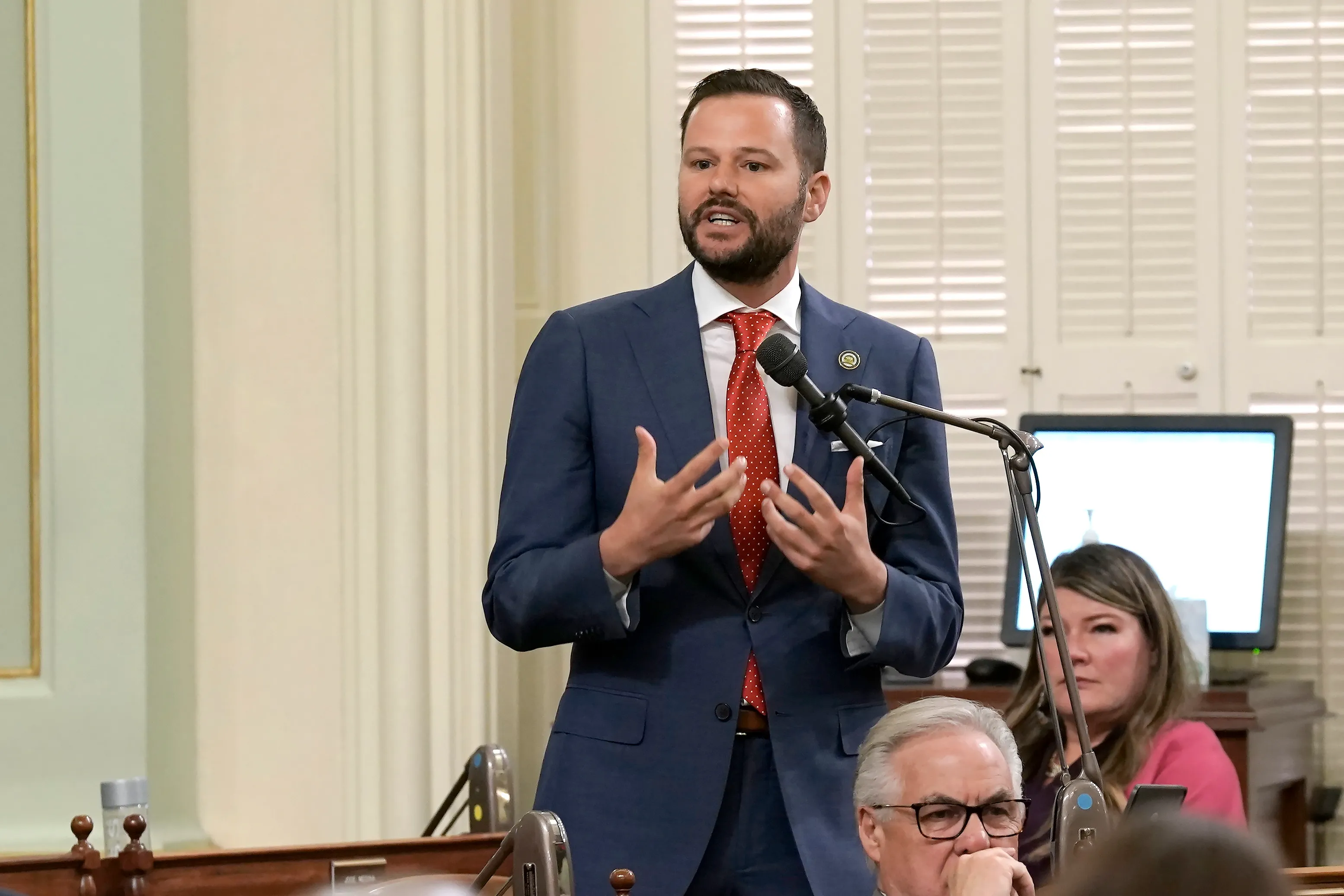 This is a photograph taken in what appears to be a formal government chamber or legislative setting. A man in a navy blue suit with a red tie is standing and speaking at a microphone, gesturing with his hands in an animated manner typical of political speech. The setting features elegant cream-colored curtains, white shuttered windows, and formal wooden furnishings that suggest this is likely a state capitol or similar government building. In the background, other individuals are seated, incl...