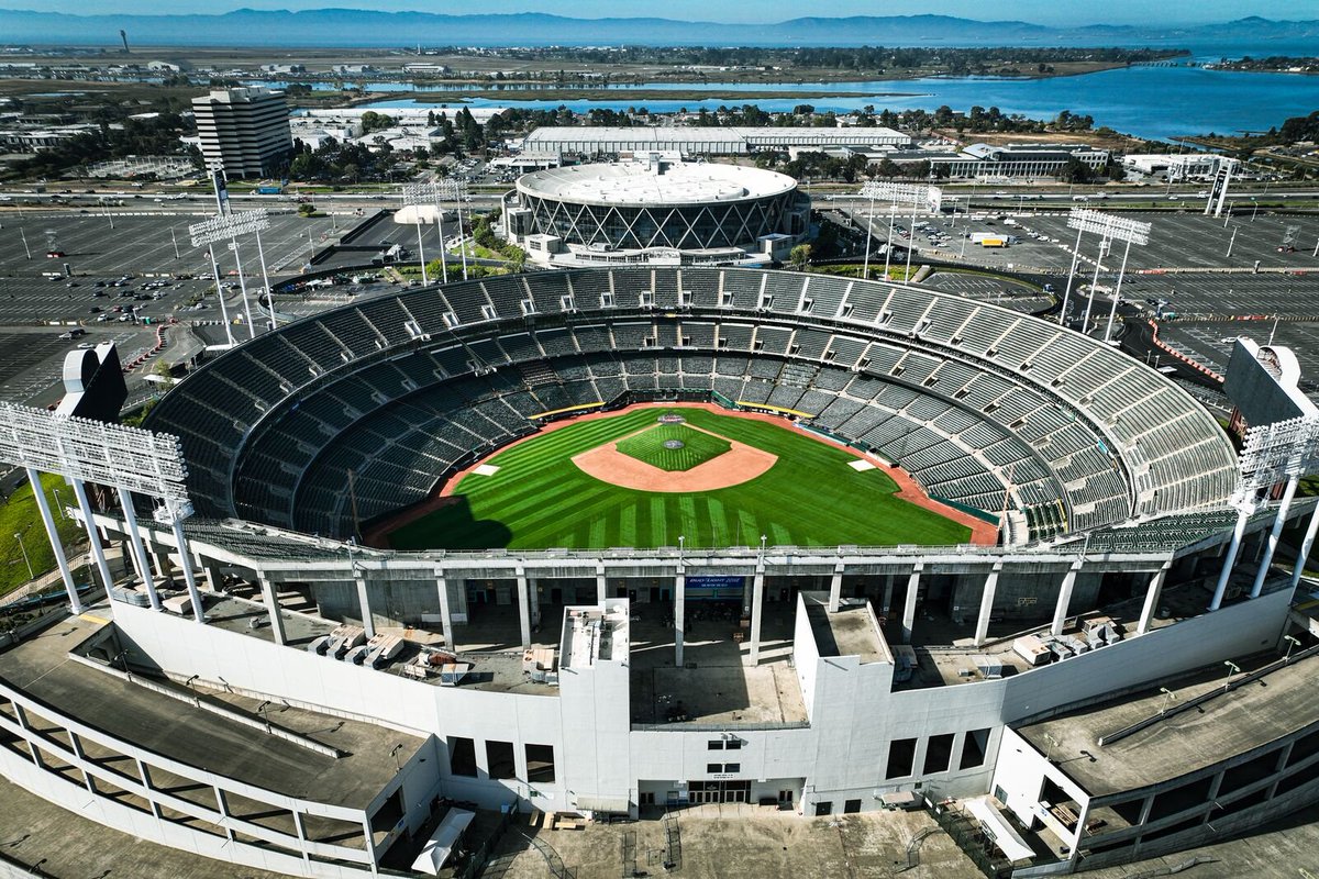 This is an aerial photograph showing a large baseball stadium taken from a high vantage point. The image captures a classic multi-tiered baseball stadium with distinctive white concrete architecture and multiple levels of seating arranged in a circular pattern around a pristine baseball diamond. The field features vibrant green grass with characteristic diamond-shaped dirt infield and pitcher's mound clearly visible. In the background, there's a smaller circular white building and a body of w...