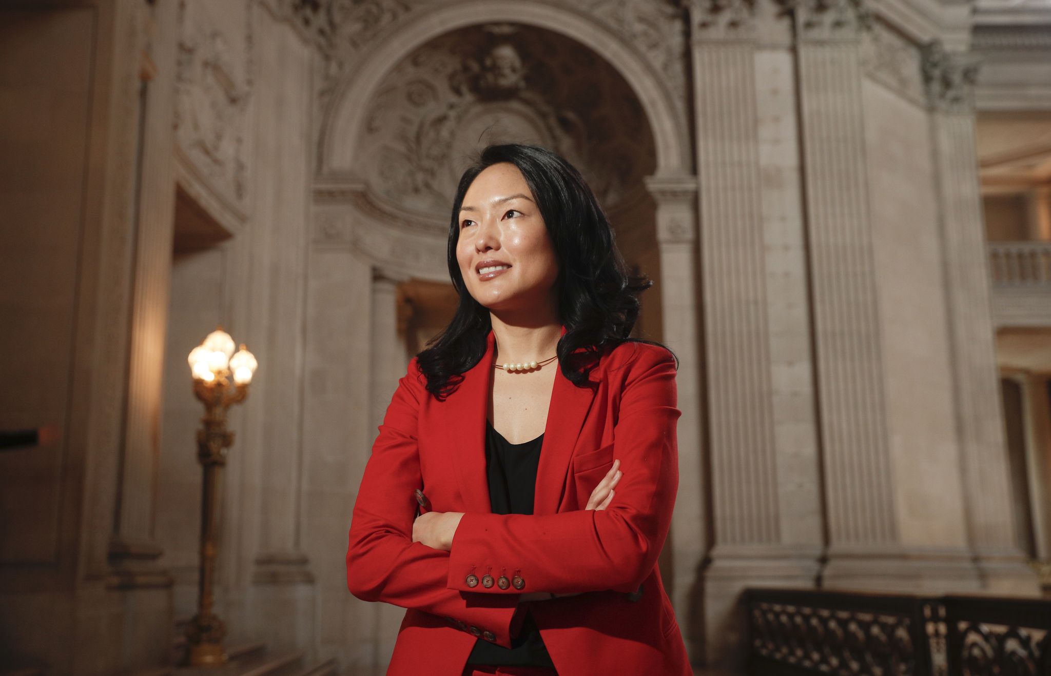 This is a professional portrait photograph of a woman in formal business attire standing in what appears to be an ornate government building or similar institutional setting. She is wearing a bright red blazer over a black top with a pearl necklace, and has her arms crossed in a confident pose while looking upward with a slight smile. The background features classical architecture with marble columns, ornate carved details, and warm lighting from traditional fixtures. The formal governmental ...