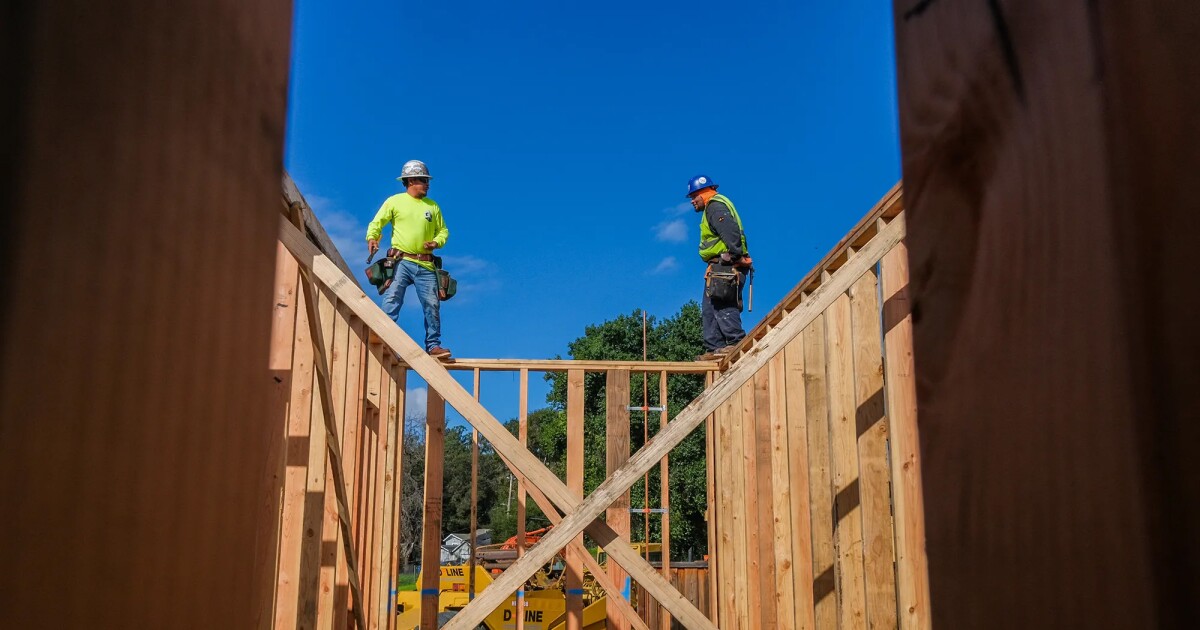 This is a photograph showing two construction workers in bright green high-visibility safety shirts and hard hats working on the wooden frame structure of a house under construction. The image is taken from below looking up, creating a dramatic perspective against a bright blue sky with some trees visible in the background. The workers are positioned on top of the wooden wall frames, which appear to be made of light-colored lumber in the typical stick-frame construction style. The composition...