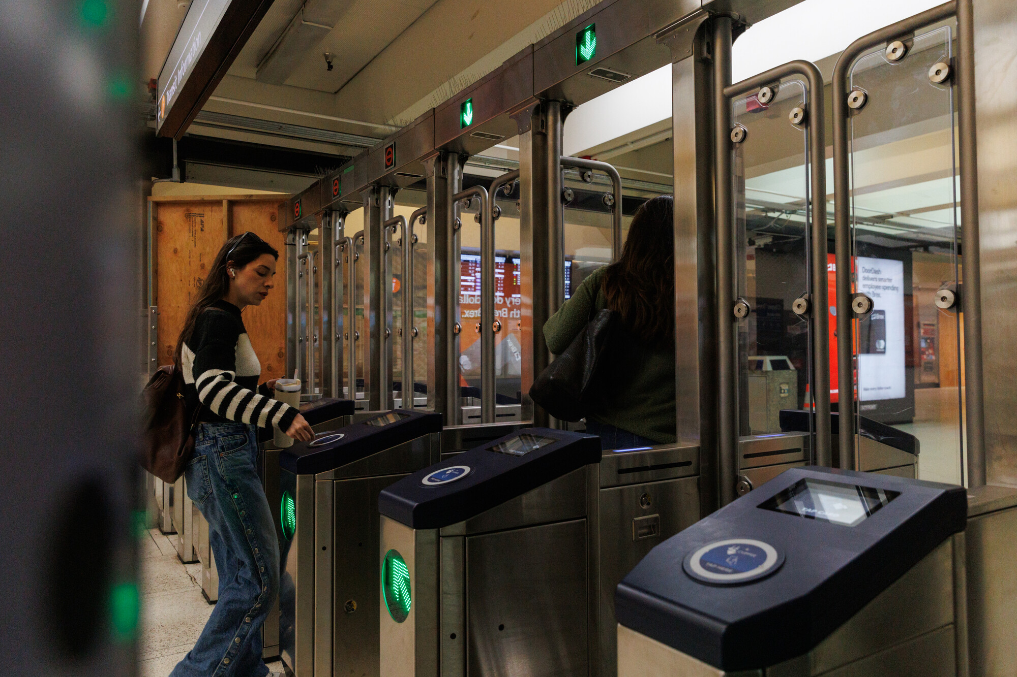 This is a photograph showing the interior of what appears to be a modern public transit station or metro system. The image captures turnstiles or fare gates with sleek metallic and glass construction. Two people are visible - one woman on the left wearing a striped sweater and jeans who appears to be using or approaching a fare gate, and another person in dark clothing on the right. The turnstiles feature black scanning surfaces with circular blue elements, likely for contactless payment or c...