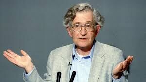 This is a photograph of an elderly man who appears to be in his 70s or 80s, speaking or presenting in what looks like a lecture or talk setting. He has white/gray curly hair and is wearing a light gray blazer over a collared shirt. He is gesturing with both hands open and raised slightly, in an expressive speaking pose, suggesting he is making a point or explanation. The background is a blurred dark/neutral tone, keeping focus on the subject. The image has a candid, documentary-style quality ...