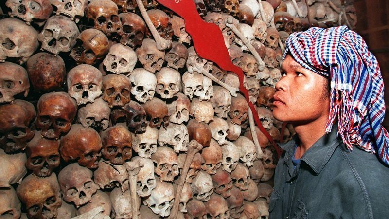 This is a photograph depicting a person wearing a traditional Cambodian krama (checkered scarf) on their head, standing in front of a large memorial stupa filled with human skulls and bones. The wall behind them is densely packed with hundreds of human skulls and long bones, stacked from floor to ceiling. A red flag or banner is visible among the bones. The scene is strongly associated with the Killing Fields of Cambodia, specifically the Choeung Ek genocidal center near Phnom Penh, where vic...