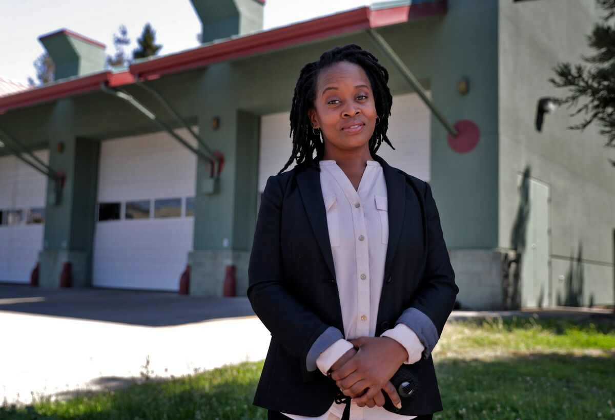 This is a professional photograph of a woman standing outdoors in front of what appears to be a fire station, identifiable by its large white garage doors, green and red architectural trim, and fire hydrants visible in the background. The woman has black locs hairstyle, is wearing a black blazer over a light pink/white button-up blouse, and is holding what appears to be a face mask in her clasped hands. She also wears a smartwatch. The image has a professional, portrait-style composition with...