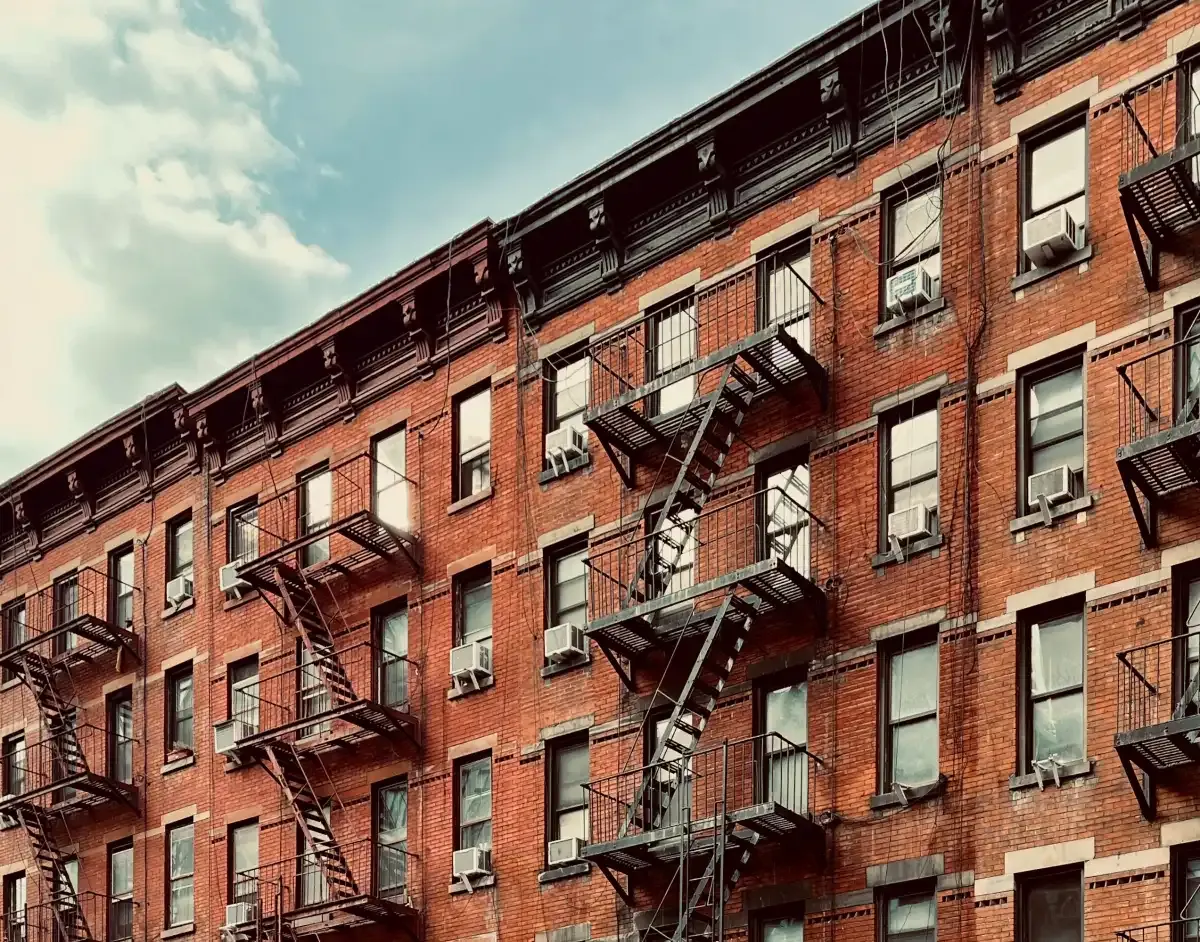 This is a photograph showing a classic red brick apartment building with characteristic New York City tenement architecture. The image captures multiple stories of the building shot from a low angle against a cloudy blue sky. The facade features numerous windows with air conditioning units, black metal fire escapes zigzagging down the exterior, and ornate decorative cornice work at the roofline. The brick construction appears weathered and aged, typical of early 20th century urban housing. Th...