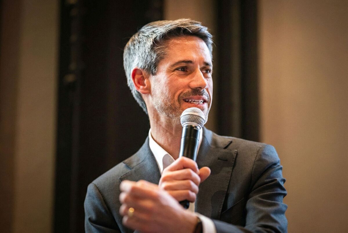 This is a professional photograph of a man speaking at what appears to be a formal event or conference. The subject is a middle-aged man with salt-and-pepper hair and light stubble, wearing a dark gray suit jacket with a white dress shirt open at the collar. He is holding a handheld microphone close to his mouth and appears to be mid-speech, with a slight smile on his face. His other hand is raised in a gesturing position, suggesting he is actively engaging with an audience. He is also wearin...