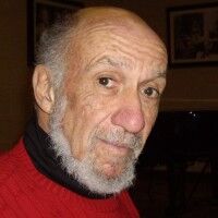 This is a casual photographic portrait of an elderly man, likely taken indoors in a home or restaurant setting. The man has a white/gray beard and is wearing a red shirt or jacket. He appears to be in his 70s or older, with a serious or contemplative expression. The background is slightly blurred but shows warm indoor lighting and what appears to be framed artwork or pictures on the wall. The photo has an informal, snapshot quality suggesting it was taken at a social gathering or family event...