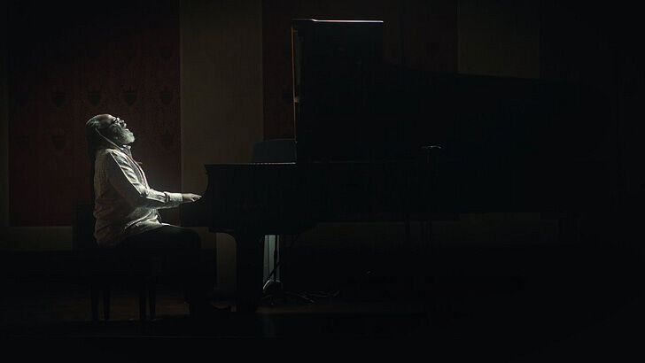 This is a moody, artistic photograph showing a person playing piano in dramatic low lighting. The image is composed in a cinematic style with strong contrast between light and shadow, creating an atmospheric and contemplative mood. The pianist is seated at what appears to be a grand piano, captured in profile with their head tilted back slightly, suggesting deep emotional engagement with the music. The lighting creates a spotlight effect on the musician while leaving much of the surrounding s...