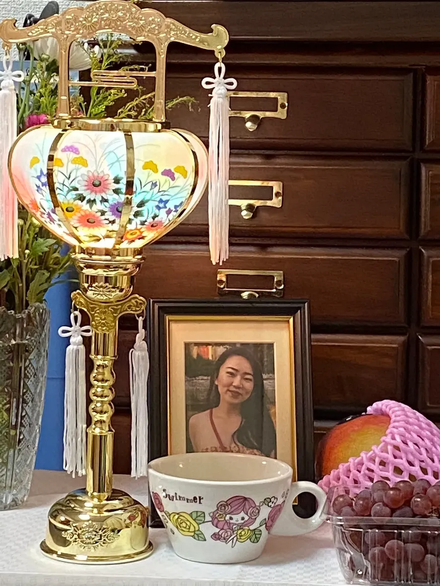 This is a photograph showing what appears to be a memorial or tribute display arranged on a surface. The centerpiece is an ornate golden Chinese lantern with floral patterns and decorative tassels. In front of it sits a framed photograph of a young Asian woman who is smiling at the camera. The arrangement includes a decorative teacup with cartoon characters, a glass bowl containing what appears to be nuts or dried fruit, fresh flowers in a vase, and wooden furniture in the background. The ove...