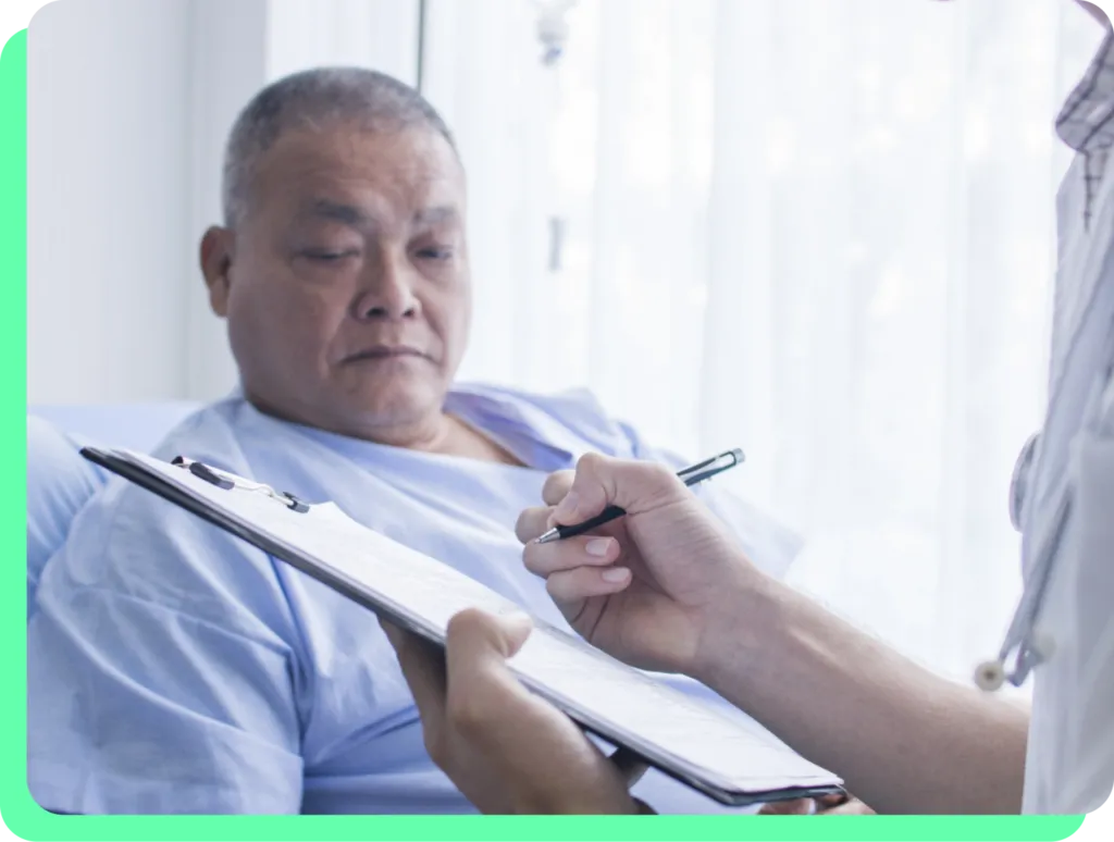 This is a photograph showing a healthcare consultation scene in what appears to be a hospital or medical facility. The image shows an older Asian man wearing a light blue hospital gown lying in a hospital bed, looking somewhat concerned or tired. In the foreground, we can see the hands and partial white coat sleeve of a healthcare provider who is holding a clipboard with papers and a pen, appearing to be taking notes or reviewing medical information. The setting has a clean, clinical appearan...