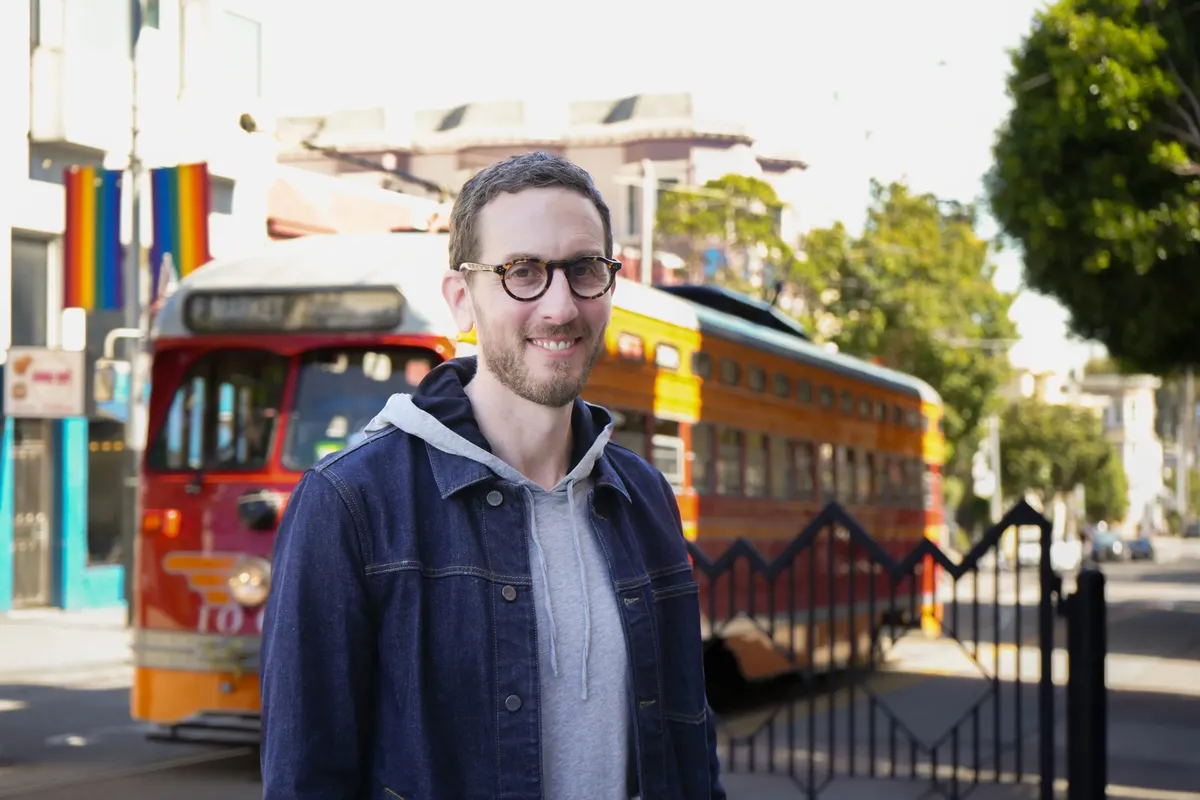 This is a casual outdoor photograph showing a smiling man with glasses, beard, and dark hair wearing a navy denim jacket over a gray hoodie. He's standing on what appears to be a city street or transit area, with colorful buses visible in the background - one appears to be red and yellow/orange. There are also rainbow pride flags visible on buildings in the background, suggesting this may be in or near a LGBTQ-friendly district. The setting has an urban, public transportation feel with metal ...