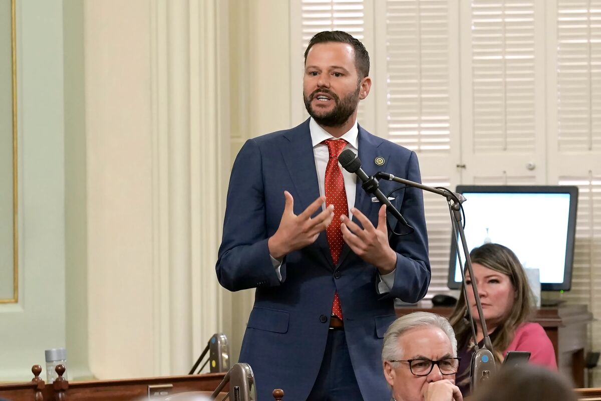 This is a photograph taken in what appears to be a formal government chamber or legislative setting. A man in a navy blue suit with a red tie is standing and speaking at a microphone, gesturing with his hands in an animated manner typical of political speech. The setting features elegant cream-colored curtains, white shuttered windows, and formal wooden furnishings that suggest this is likely a state capitol or similar government building. In the background, other individuals are seated, incl...
