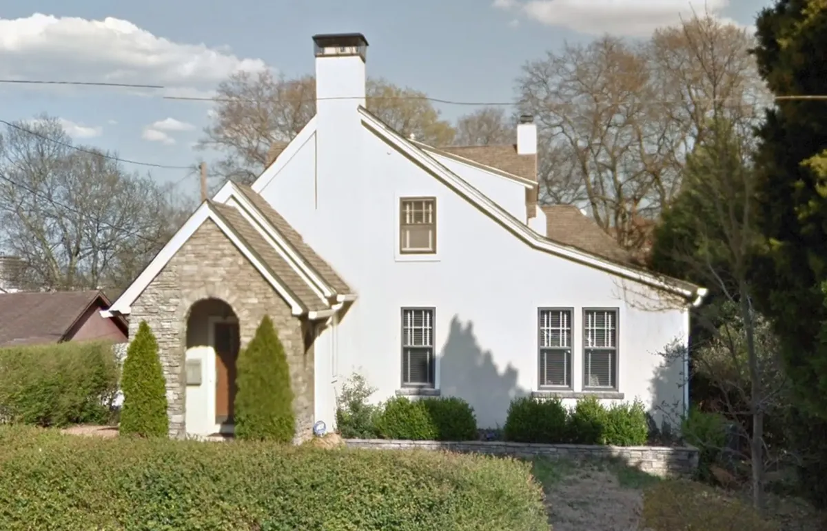 This is a photograph of a charming single-story residential house featuring a distinctive architectural style. The home has white stucco or painted walls with a natural stone accent section around the arched front entrance, creating an appealing contrast in textures and colors. The house features a steep-pitched roof with multiple gables and a prominent white chimney. The front facade includes several multi-paned windows with white trim that complement the overall aesthetic. Well-maintained l...