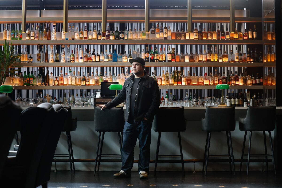This is a professional photograph of a man standing behind the bar of an upscale establishment. The image showcases an impressive wall-to-wall display of spirits and liquors arranged on multiple shelves, creating a warm, amber-lit backdrop that suggests this is a high-end whiskey bar or cocktail lounge. The man, wearing a black beret, dark button-up shirt, jeans, and casual shoes, appears to be the bartender or owner, positioned confidently with his hands on the bar counter. The foreground fe...