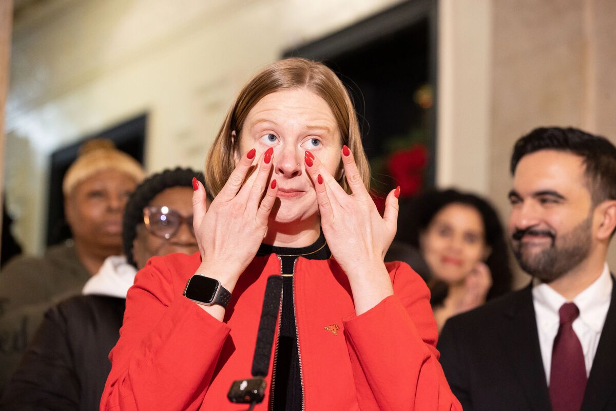 This is a photograph showing a woman in a red blazer with her hands covering her face in what appears to be an emotional reaction, possibly crying or showing distress. She has red nail polish and is wearing a smartwatch. The woman appears to be at some kind of formal event or press conference, with several people visible in the background including a man in a suit with a burgundy tie who appears to be smiling. The setting looks like an indoor venue with blurred architectural elements visible....