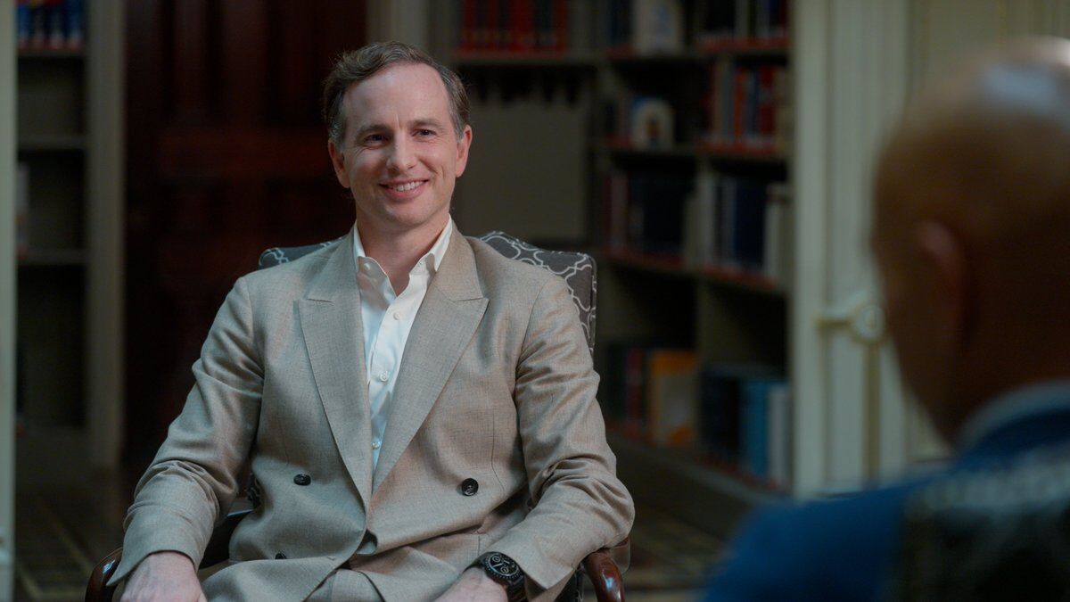This is a professional photograph showing a man in business attire sitting in what appears to be an office or study setting. He's wearing a light-colored double-breasted suit jacket with a white dress shirt and has a warm, confident smile. The setting features bookshelves filled with books in the background, suggesting an academic or professional environment. The man is seated in what looks like an ornate chair and is wearing what appears to be a luxury watch. The lighting and composition are...