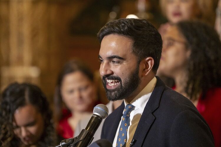 This is a color photograph taken at what appears to be a press conference or public speaking event. The main subject is a young man with dark hair and a beard, smiling broadly while speaking into a handheld microphone. He is wearing a navy blue blazer, white dress shirt, and a blue tie with a gold/yellow dot pattern. The setting appears to be an ornate indoor venue with warm wood-toned walls visible in the background. Several other people are blurred in the background, including women in red ...