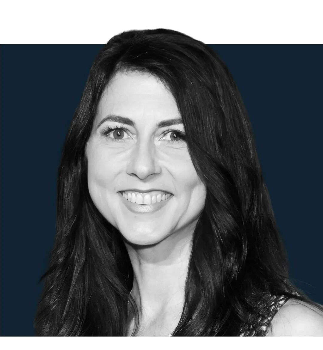 This is a professional black and white portrait photograph showing a woman with long, wavy dark hair smiling at the camera. She appears to be wearing a sleeveless top with a textured or patterned fabric. The image has a formal, polished quality typical of professional headshots or red carpet photography, with a dark blue background. The black and white treatment gives it a classic, timeless aesthetic. The woman's pose and expression suggest this was taken at a formal event or professional pho...