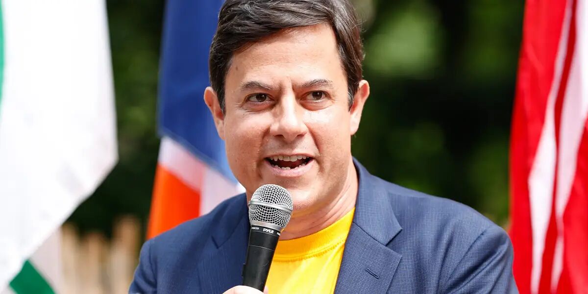 This is a color photograph of a middle-aged man with dark brown hair speaking into a Pyle-brand handheld microphone at what appears to be an outdoor public event or rally. He is wearing a bright yellow t-shirt underneath a navy blue blazer, suggesting a semi-casual public appearance. Behind him, several flags are visible — including what appears to be a blue and orange flag, an American flag (red and white stripes visible), and what may be an Indian flag (green and white visible) — indicating...