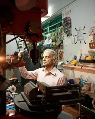 This is a color photograph depicting an older man, likely in his 60s or 70s, working in what appears to be a small workshop or repair shop. He is wearing a light pink button-down shirt and is reaching upward to operate what looks like a drill press or milling machine. The workshop background is cluttered and characterful, with a pegboard wall covered in various tools, notes, and decorative items including what appears to be a stuffed animal or doll. The workbench area is filled with machinery...