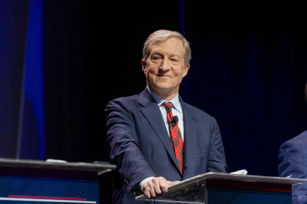 This is a professional photograph taken at what appears to be a political debate or formal event. The image shows an older man with light gray-blonde hair, wearing a navy blue suit with a light blue dress shirt and a red plaid/tartan tie. He is standing at a clear podium/lectern and appears to be smiling or smirking confidently toward the camera or audience. A small lavalier microphone is clipped to his tie. The background features dark tones with blue accent lighting, typical of a debate or ...