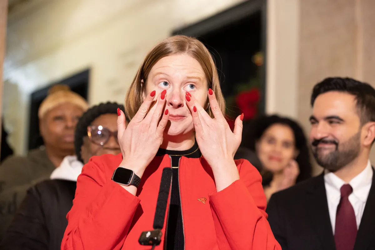 This is a photograph showing a woman in a red blazer with her hands covering her face in what appears to be an emotional reaction, possibly crying or showing distress. She has red nail polish and is wearing a smartwatch. The woman appears to be at some kind of formal event or press conference, with several people visible in the background including a man in a suit with a burgundy tie who appears to be smiling. The setting looks like an indoor venue with blurred architectural elements visible....
