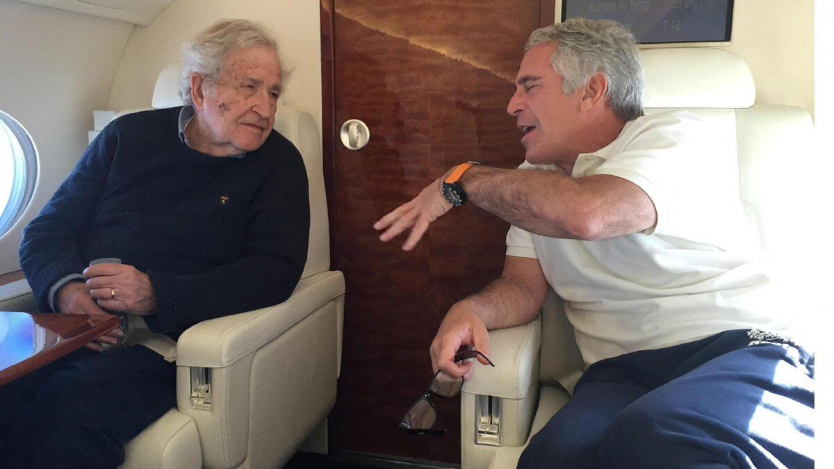 This is a candid photograph taken inside what appears to be a private jet or luxury aircraft, based on the cream leather seats, wood-paneled door, and oval window visible in the background. Two older men are seated across from each other in reclining chairs, engaged in animated conversation. The man on the left is elderly with white hair, wearing a dark navy sweater, and appears to be listening attentively. The man on the right has gray hair, is wearing a white polo shirt and dark pants, and ...