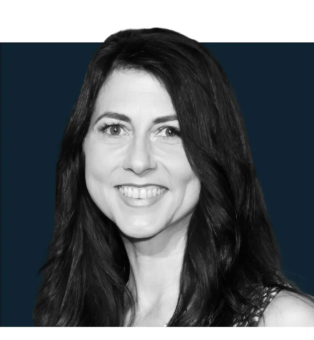 This is a professional black and white portrait photograph showing a woman with long, wavy dark hair smiling at the camera. She appears to be wearing a sleeveless top with a textured or patterned fabric. The image has a formal, polished quality typical of professional headshots or red carpet photography, with a dark blue background. The black and white treatment gives it a classic, timeless aesthetic. The woman's pose and expression suggest this was taken at a formal event or professional pho...