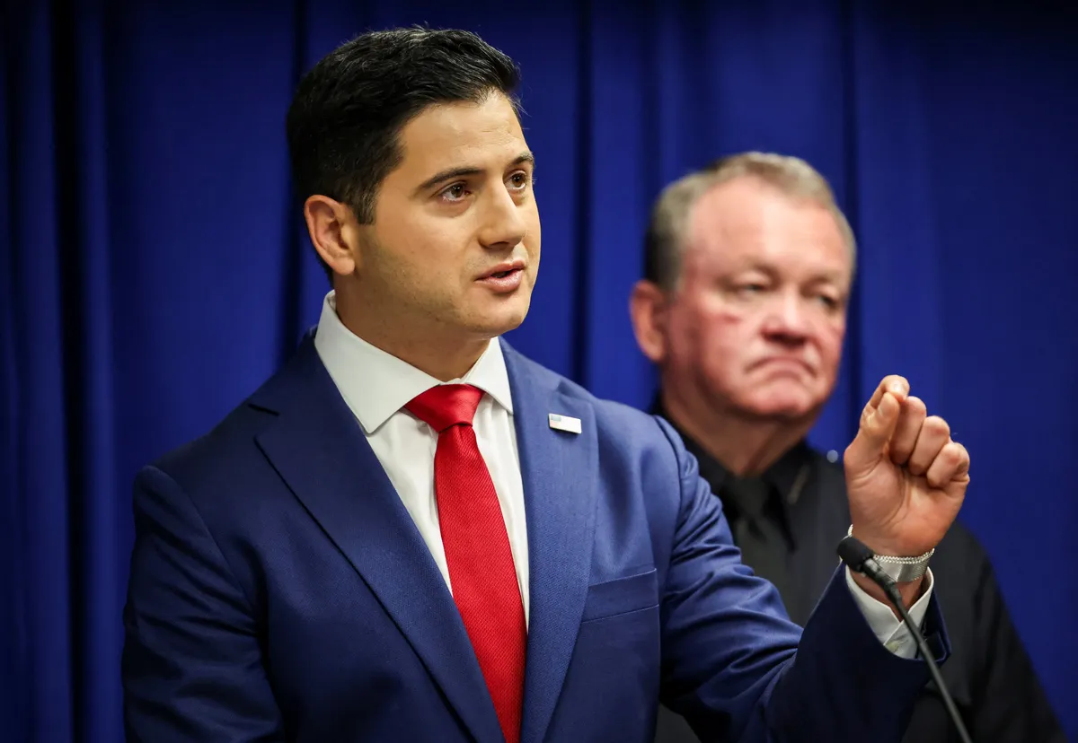 This is a professional photograph taken at what appears to be an official government press conference or announcement. The image shows a man in a navy blue suit with a red tie speaking at a podium, with his hand raised in a gesturing motion. Behind him stands another man in dark clothing against a backdrop of blue curtains, which is typical of formal government settings. The lighting and composition suggest this is an official political event or press briefing. The formal attire, official set...