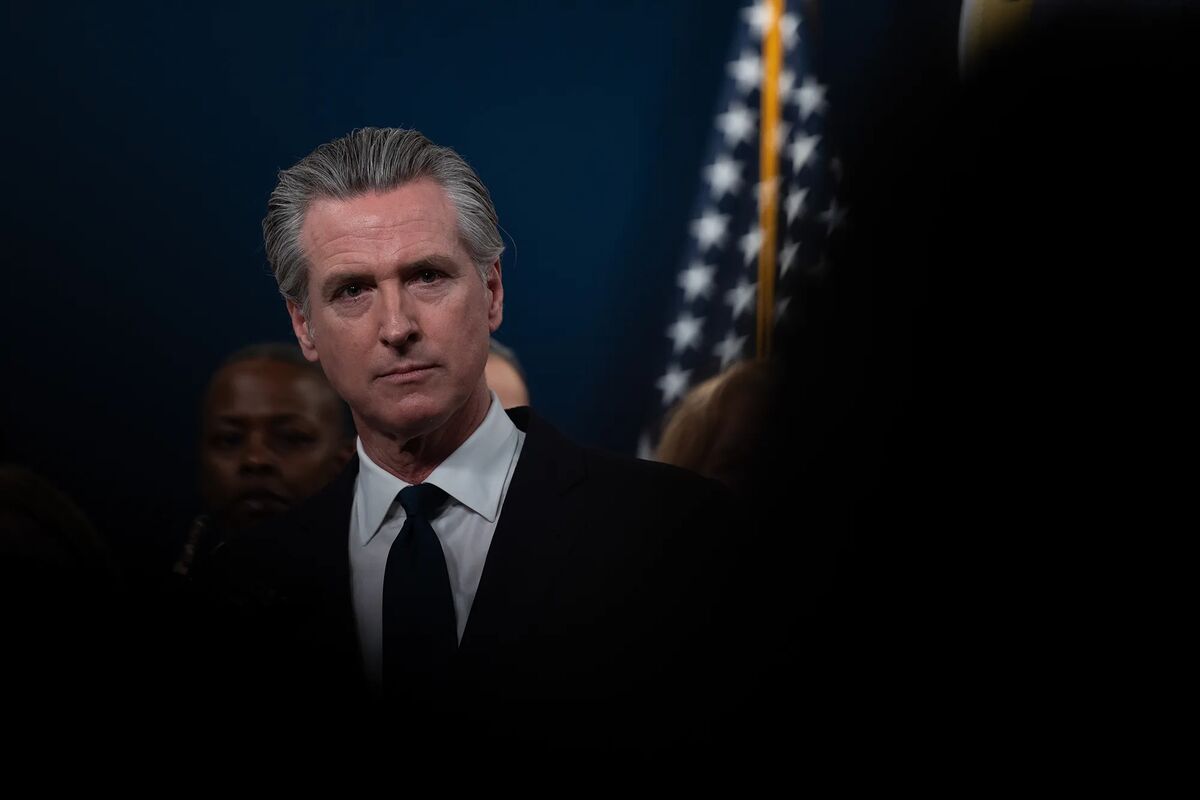 This is a professional photograph showing a man in formal attire (dark suit and black tie) at what appears to be an official government event or press conference. The image has a serious, formal tone with dramatic lighting against a dark blue background. An American flag is visible in the background, suggesting this is taking place in an official government setting. The man has graying hair and appears to be middle-aged, looking directly at the camera with a somber expression. Other figures a...